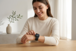 Una fotografía de estilo e-commerce profesional muestra a una mujer sonriente sentada a una mesa de madera clara, interactuando de forma natural con el Apple Watch Serie 11 46mm GPS - Negro en su muñeca izquierda. El reloj mantiene el diseño exacto de la imagen de referencia, con su caja negra y correa deportiva a juego, y la pantalla muestra la esfera analógica específica. La escena es luminosa, moderna y minimalista, con un fondo desenfocado que incluye un jarrón con una rama de olivo y una ventana con cortinas, iluminada por una luz de estudio suave y realista.