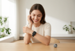 Una fotografía de estilo e-commerce profesional muestra a una mujer joven y sonriente sentada a una mesa de madera en un interior moderno y luminoso. Lleva el Oppo Watch S 4G negro en su muñeca izquierda, el cual mantiene el diseño exacto y la interfaz de la imagen de referencia. Su mano derecha reposa suavemente cerca de su rostro mientras mira el reloj con alegría. Sobre la mesa, a la derecha, se encuentra la correa gris de regalo enrollada. El fondo es minimalista y desen focado, con plantas verdes y luz natural suave que entra por una ventana, creando un ambiente de alta gama.