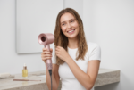 Fotografía de estilo de vida profesional y luminosa en 4K que muestra a una mujer sonriente con el cabello húmedo secándose con el secador para el pelo Dyson Nural HD16 Cera – Rosa. La mujer, vestida con una camiseta blanca, sostiene el dispositivo rosa metálico en su mano derecha mientras usa la izquierda para separar su cabello. El secador mantiene su diseño exacto con el cab ezal hueco y los botones visibles. La escena transcurre en un baño moderno y minimalista con paredes blancas, un espejo grande y una encimera de má rmol donde descansan un peine y una botella de aceite. La iluminación de estudio suave resalta los tonos de piel naturales y el acabado del producto.