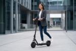 Una fotografía de estilo e-commerce profesional y de alta gama muestra a una mujer joven y sonriente, de aspecto natural y feliz, montando el patinete eléctrico Xiaomi Electric Mi Elite - Negro. El patinete, que mantiene su diseño exact o, color negro y marca visible en el mástil, está en movimiento sobre un camino de hormigón pulido en un entorno urbano moderno y minimalista con arquitectura de vidrio y acero. La mujer, vestida de forma elegante y casual con una chaqueta gris y vaqueros, tiene una mano en el manillar y mira hacia adelante con alegría. La iluminación es suave y de estudio, y el patinete está perfectamente enfocado, con un sutil desenfoque de fondo.
