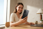 Una fotografía de estilo de vida profesional muestra a una mujer sonriente sentada a una mesa de madera, sosteniendo y mirando el teléfono inteligente Xiaomi Redmi 15 5G en color negro. El dispositivo , que coincide con la imagen de referencia, está en primer plano y en foco, mostrando su diseño y el módulo de cámara trasera. La mujer viste una prenda de punto beige y tiene una expresión feliz. Al fondo, hay una ventana con luz natural, una lámpara de mesa y un ordenador portátil sobre la mesa, creando un ambiente moderno y minimalista. La iluminación es suave y realista.